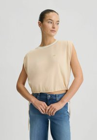 Top cropped beige realizzato in tessuto morbido, con scollatura rotonda e maniche arrotolate. Presenta un dettaglio del logo dorato sul davanti e cordini all'orlo.