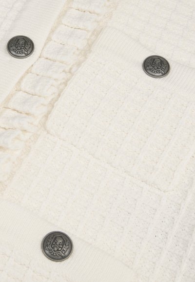 Pull en tricot écru avec des motifs texturés, présentant un col côtelé, une poche et des boutons en métal foncé avec un design de crâne.