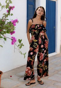 Blommig jumpsuit med svart bakgrund, prydd med volanger och vida ben, dekorerad med röda och rosa blommor. Bärs med svarta flip-flop sandaler.