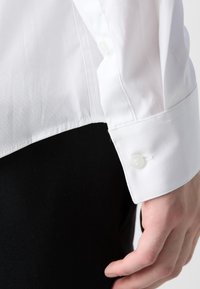 Rechterhand dicht bij de heup, draagt een wit overhemd met lange mouwen en knopenmanchet, zwarte broek zichtbaar onder de shirtzoom.