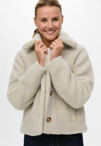 Veste en polaire couleur crème avec un col, des boutons devant et une surface texturée. Présente une coupe ample et est portée sur un haut blanc.