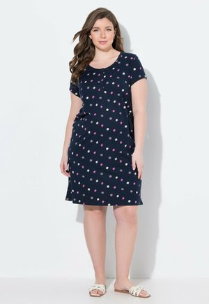 Mujer de pie con un vestido azul marino de manga corta con estampados florales rosas y blancos y sandalias blancas de tira, sobre un fondo sencillo.