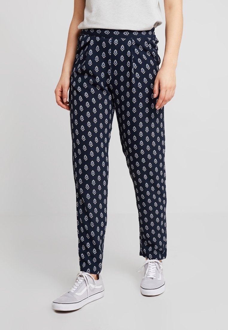 Tmavě modré kalhoty s bílými geometrickými vzory, vyrobené z lehké látky. Mají pohodlný střih a zužující se nohavice, doplněné bílými teniskami.