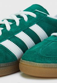 Gröna mockaskor har vita zigzag-randiga detaljer, vita snörning och en texturerad gummisula. Närbild framhäver detaljerna.