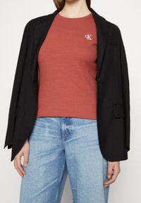 Černý blejzr přes rezavě zbarvený ribbed top s malým bílým logem "cK". Světle modré džíny se zesvětlovanou texturou.