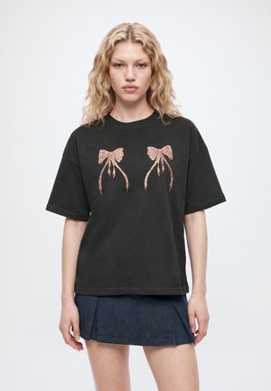 Un mannequin aux cheveux blonds ondulés porte un T-shirt noir orné de deux motifs de nœuds roses et une mini-jupe plissée foncée, sur un fond de studio clair.