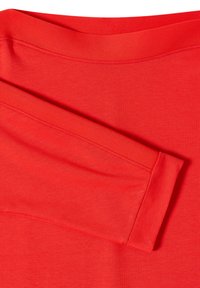 Punainen puuvillakangas, jossa on sileä pinta, pyöreä kaula-aukko ja lyhyet hihat. Huomattavat ompeluyksityiskohdat reunoilla.