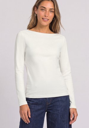 Femme portant un haut blanc à manches longues et un jean bleu, debout devant un fond clair uni, souriant doucement.