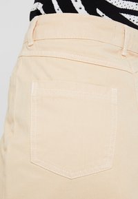 Pantalones de mezclilla beige claro con un bolsillo trasero, con costuras tonales y un acabado ligeramente texturizado. La cinturilla está asegurada con un botón.