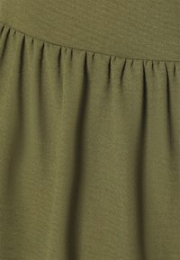 Oliivinvihreä kangas, jossa on teksturoitu pinta; ominaisuuksiin kuuluu kerrottu vyötärö, jossa on tasaisesti sijoitettuja laskoksia ja pehmeä, valuva drapeeraus.