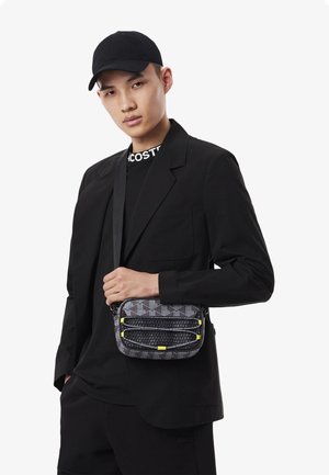 Black shoulder bag featuring a geometric pattern with textured accents and yellow zippers, carried by an individual in a black outfit.