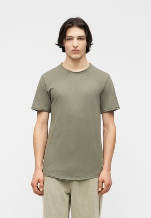 Ung mann med mellomlangt mørkt hår, iført en olivengrønn kortermet t-skjorte med rund hals og lyse beige bukser, stående mot en ensfarget bakgrunn.