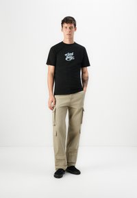 Zwarte katoenen t-shirt met een lichtblauwe grafische print, in combinatie met kaki cargobroek met meerdere zakken en een ontspannen pasvorm. Zwarte schoenen.