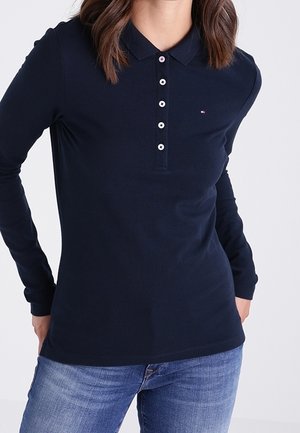 Femme portant un polo à manches longues bleu marine foncé avec des boutons blancs et un jean bleu, debout les mains derrière le dos.