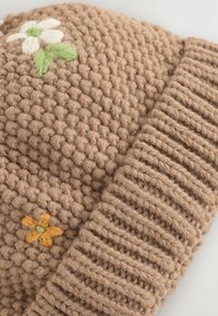 Strickmütze in Beige mit strukturiertem Muster, geripptem Rand und gestickten Blumenmotiven in Weiß, Grün und Orange.