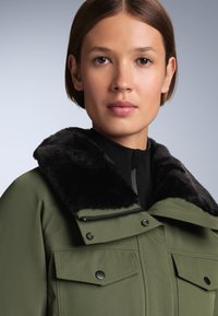 Olivno zelena jakna s črnim umetnim krznom okoli ovratnika, z zadrgo in dvema prednjima žepoma s pritisnimi gumbi, gladka tkanina in strukturirana zasnova.
