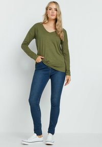 Olijfgroene lange mouwen top met een V-hals, gecombineerd met donkerblauwe skinny jeans en witte sneakers. De top heeft een relaxte pasvorm en een zijzak.