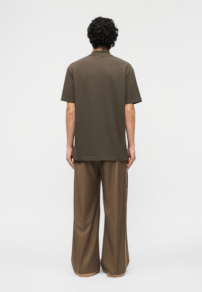 Άνδρας με σγουρά σκούρα μαλλιά στέκεται με την πλάτη, φοράει σκούρο καφέ κοντομάνικο πουκάμισο και φαρδιά, καφέ παντελόνια με ανοικτά πόδια.