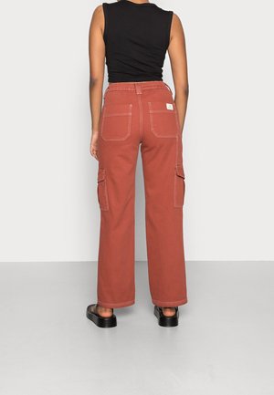 Persona vistiendo pantalones cargo de pierna ancha color óxido y una camiseta sin mangas negra, de pie de espaldas sobre un fondo liso.