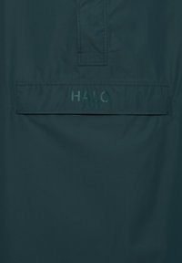 Dunkelgrüne Jacke aus glattem Stoff mit einer Klappentasche und dem "HALO"-Logo in der Mitte. Minimalistisches Design ohne sichtbare Beschläge.