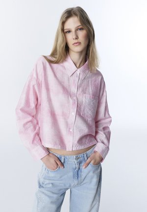 Różowa krótka koszula na guziki z lekkiego materiału z kieszonką; posiada długie rękawy i luźny krój, zestawiona z jasnoniebieskimi dżinsami.