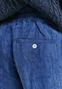 Pantaloni in lino blu con una texture stropicciata, dotati di una tasca posteriore e di un piccolo bottone bianco. Il cinturino è elasticizzato per maggiore comfort.