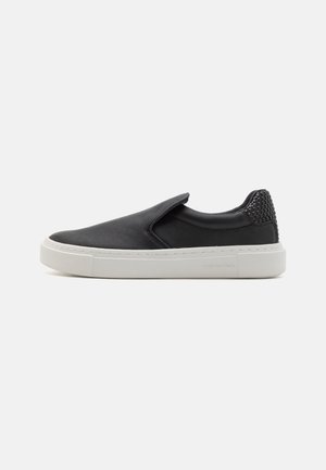 Zapatilla negra tipo slip-on con suela de goma blanca y detalle texturizado tejido en el talón, mostrada de perfil sobre un fondo blanco.