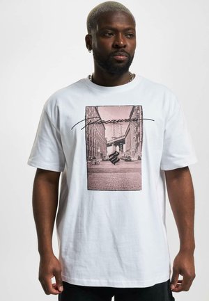 T-shirt en coton blanc avec un grand graphisme central représentant une scène urbaine noire et rose, encadrée de lignes noires et de texte stylisé.
