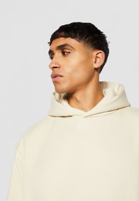 Beige hettegenser med en myk tekstur, med en hvit brodert logo på brystet og en avslappet passform.