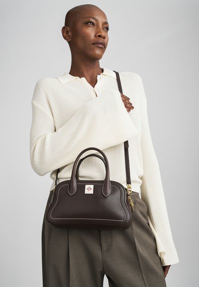 Une femme portant un pull côtelé crème et un pantalon gris tient un sac à main en cuir marron foncé avec des poignées supérieures et une bandoulière.