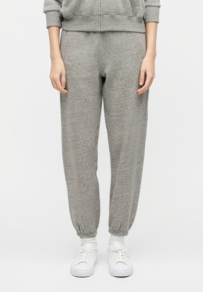 Άτομο που φοράει γκρι φόρμα με ελαστικά μανσέτες, ταιριαστό γκρι ζακέτα με φερμουάρ, λευκές κάλτσες και λευκά αθλητικά παπούτσια, όρθιο μπροστά σε λευκό φόντο.