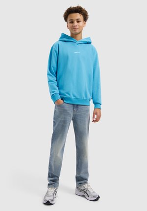 Tiener met krullend haar, gekleed in een felblauwe hoodie, verweerde blauwe jeans en witte sneakers, staand met één hand in de zak.
