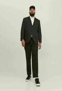 Homem com barba e cabelos em dreadlocks, vestindo um fato preto, camisa branca e ténis pretos, em pé contra um fundo claro e uniforme.