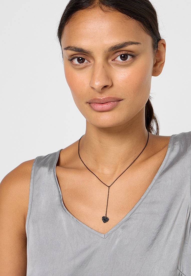 Czarny wisior w kształcie serca na delikatnym łańcuszku, noszony na szarej bluzce bez rękawów. Gładka tekstura, minimalistyczny dizajn i dyskretna elegancja.