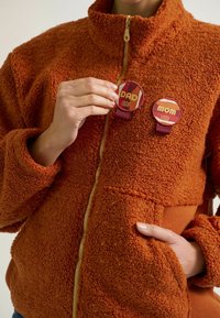 Orangene Fleecejacke mit einem hohen Kragen, ausgestattet mit einem goldenen Reißverschluss und zwei Abzeichen, versehen mit den Aufschriften "DAD" und "MOM", die an der Vorderseite befestigt sind.