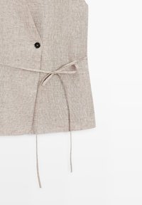 Beige linnen vest met een enkele zwarte knoop en een dunne strikceintuur geknoopt in de taille, afgebeeld tegen een witte achtergrond.