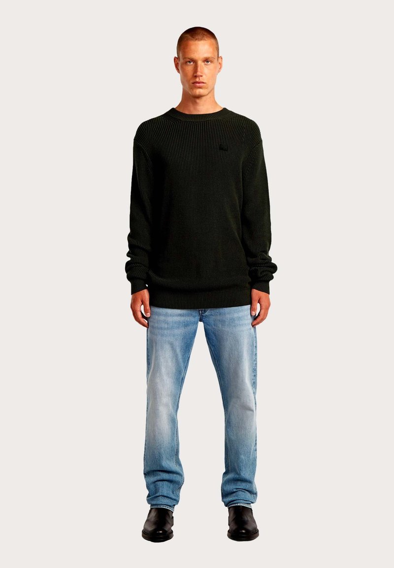 Pull tricoté vert foncé à col rond, associé à un jean bleu clair et des bottes noires. Le pull présente une texture côtelée et une poche.