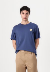 Blue cotton t-shirt with short sleeves, featuring a small yellow sun graphic on the chest, paired with beige trousers.