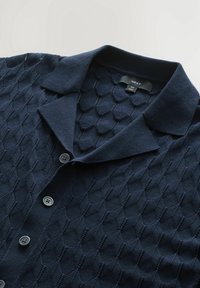 Cardigan en maille bleu marine avec motif en losange, doté d'un col, de six boutons et d'un tissu texturé. Étiquette de taille visible.