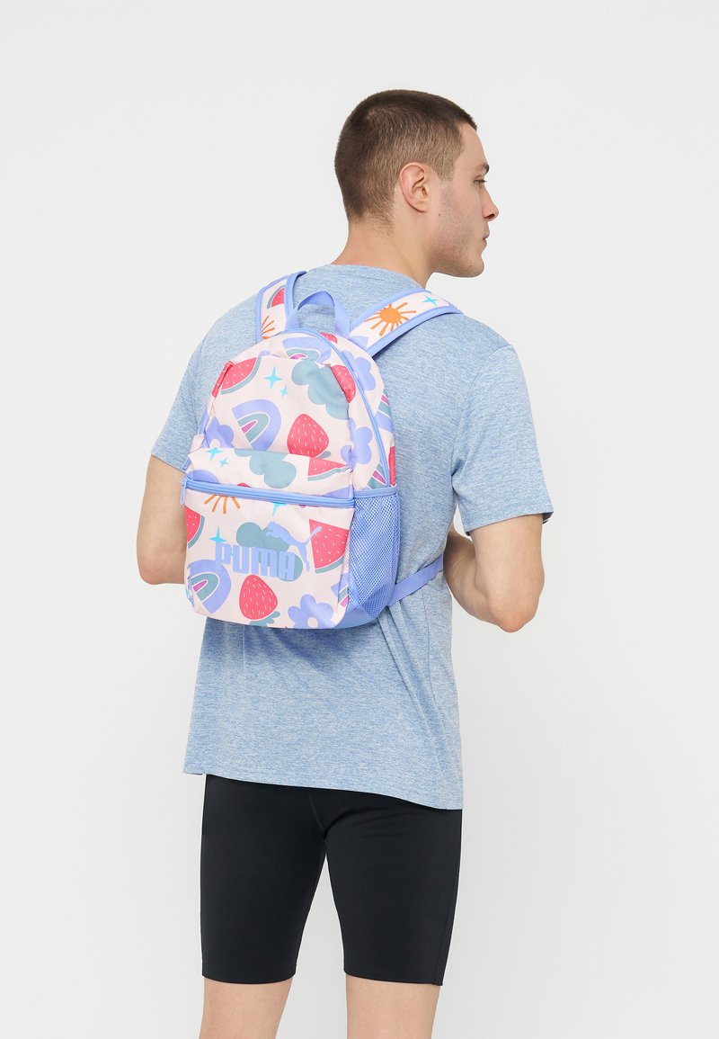 Hombre con camiseta azul claro y pantalones cortos negros con una mochila pastel con patrones coloridos de frutas y sol.