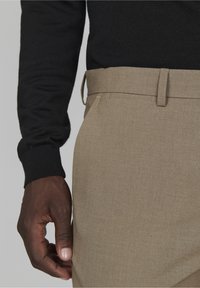 Pantalons beiges au toucher lisse, avec une coupe droite, des passants de ceinture et un motif subtil. Une main repose sur la taille.