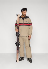 Σακίδιο σκι με μπεζ πουλόβερ με κόκκινες και μαύρες ρίγες, μπεζ παντελόνια και μαύρα μποτάκια σκι. Κρατάει ένα ζευγάρι σκι σε ένα χέρι.