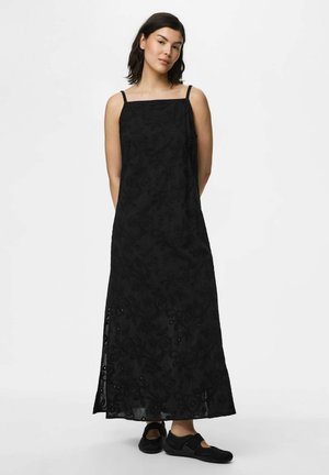 Femme portant une robe maxi noire sans manches avec un overlay en dentelle florale et une fente latérale, associée à des chaussures plates noires, debout sur un fond blanc.
