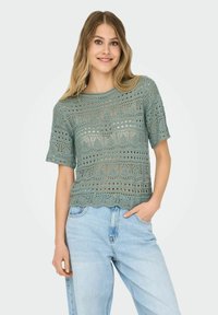 Top de ganchillo verde claro con mangas cortas, que presenta un patrón de calado y un dobladillo festoneado, combinado con jeans de denim azul claro.