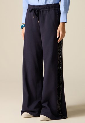 Kobieta w granatowych spodniach o szerokich nogawkach z czarnymi cekinowymi paskami po bokach, z paskiem ze sznurkiem w talii, w zestawieniu z jasnoniebieską koszulą i białymi butami.