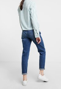 Camicia a maniche lunghe a righe in un verde acqua chiaro e bianco abbinata a jeans in denim blu arrotolati e scarpe da ginnastica bianche, vista da dietro.