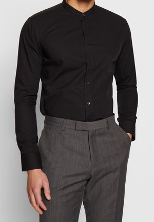 Zwart shirt met lange mouwen en een mandarin kraag, voorzien van een plooi met knopen aan de voorkant. Gecombineerd met textuurgrijze broek.