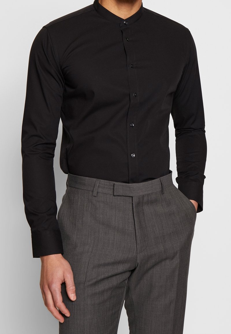 Czarna koszula z długim rękawem i kołnierzem typu Mandarin, z przodu zapinana na guziki. W połączeniu z teksturowanymi szarymi spodniami.