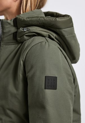 Veste isolée verte avec un col haut, une capuche à cordon de serrage et un patch logo en caoutchouc noir sur la manche. Texture lisse et design élégant.