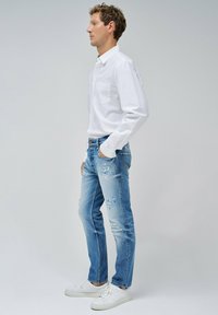 Camisa blanca de botones con bolsillo en el pecho, jeans de mezclilla desgastados de color azul claro y zapatillas blancas. Los jeans muestran desvanecimiento y pequeños desgastes.